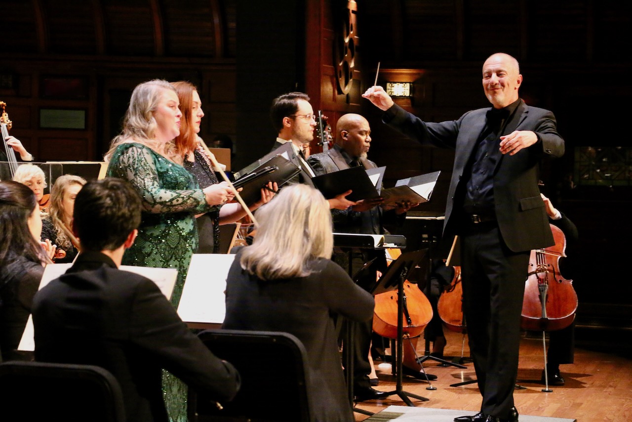 A person wearing black standing on a podium conducting four standing singers surrounded by 3 violinists, a bassist, 3 viola players, and three cellists.