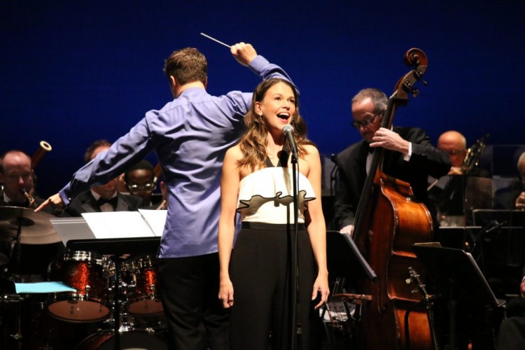 Sutton Foster singing into a mic on-stage. John Devlin conducts the orchestra behind her.