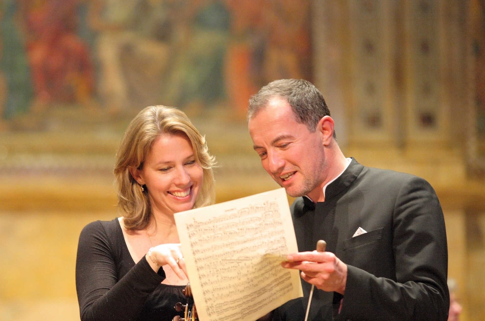 A female violinist and male conductor smile as they look over a page of music