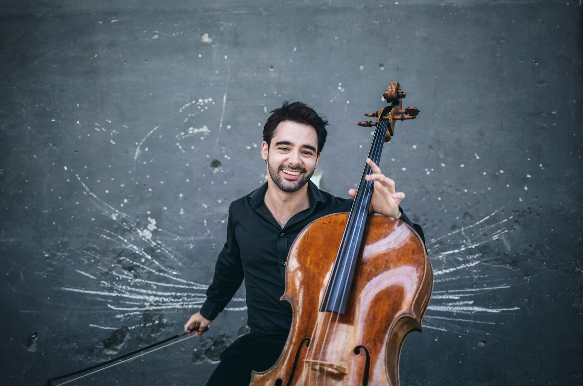 Pablo Ferrández holds his cello up in front of a spatter-patterned wall.