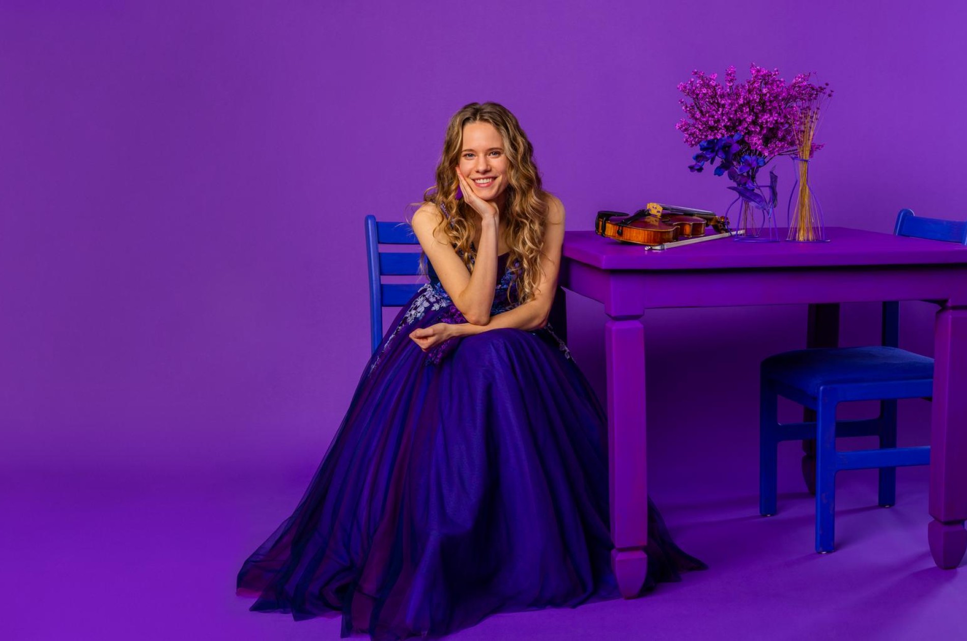 A monochromatic image of a violinist in a purple gown sitting beside her instrument upon a purple table on a purple ground beside a purple wall.
