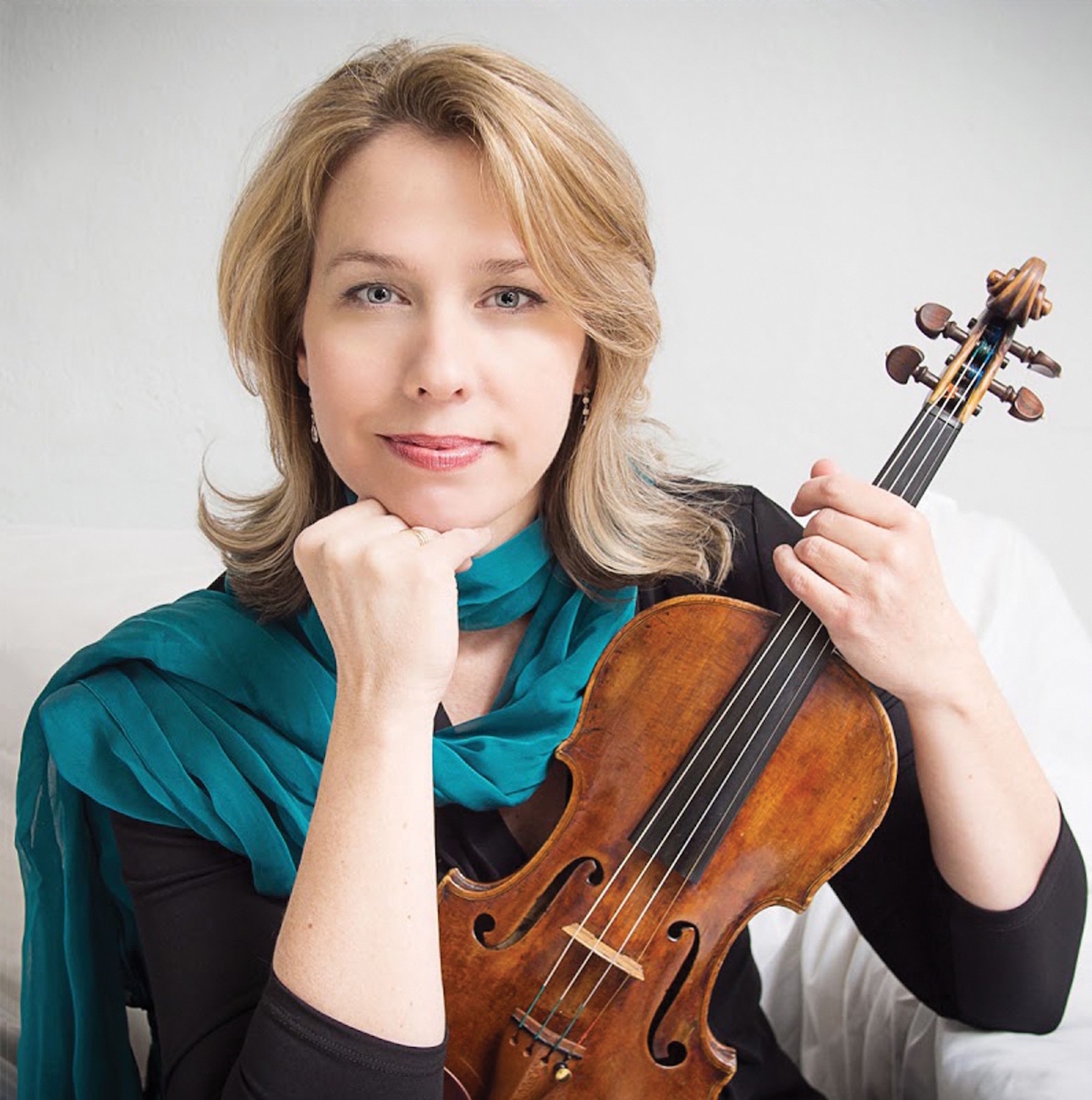 Wearing a black top and teal scarf, a blond woman holds up her violin.