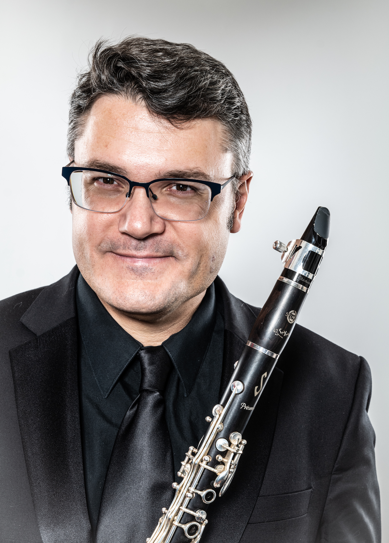 A clarinetist attired in concert black posing in front of a light gray background while holding their instrument.