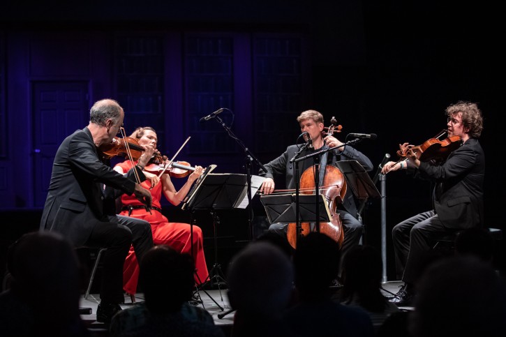 Action shot of the Signum Quartet performing. They are seated next to each other in a slight semi-circle. The backs of the audience's heads are visible in shadow at the bottom of the image.