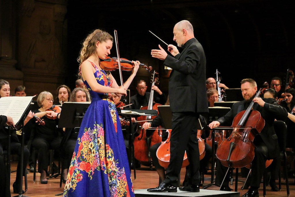 A violinist performing as soloist on stage with a conductor and 14 members of a full orchestra playing string instruments.