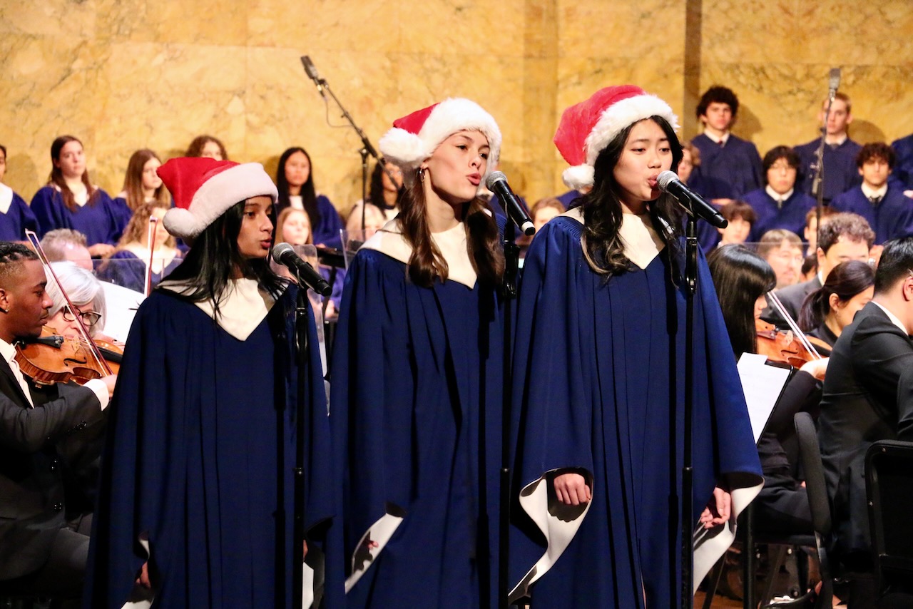 Three choir soloists in blue robes wearing Santa hats while singing in front of orchestral string musicians and other choir members onstage.