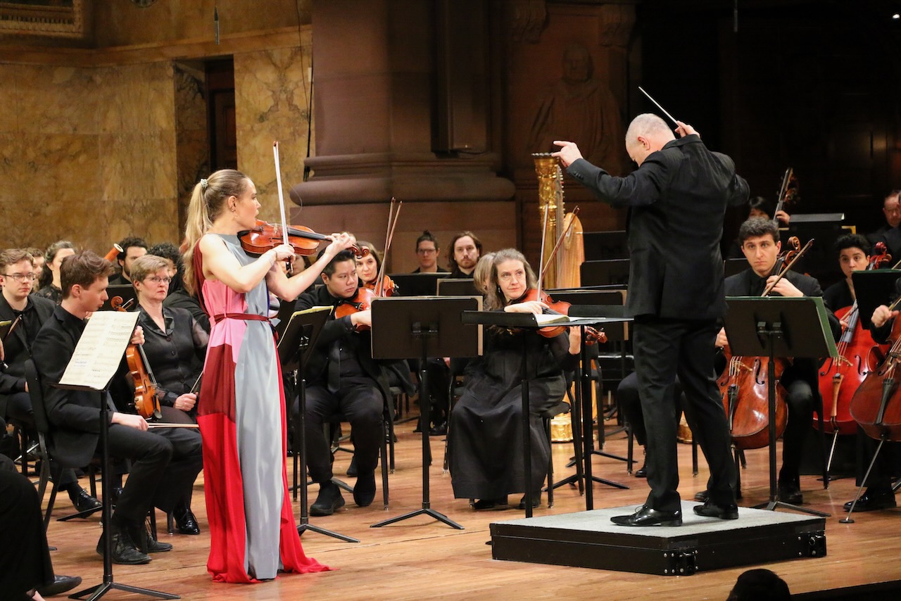 Solo violinist performing on stage with a conductor beside them leading multiple musicians including a bassonist, 6 violists, 3 cellists, 2 double bassists, and a harpist.