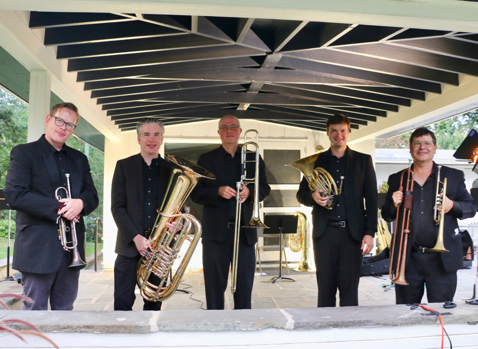 A brass quintet featuring musicians holding their instruments: trumpet, tuba, trombone, french horn, english horn/trumpet, respectively.