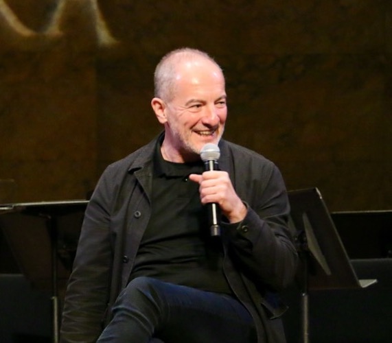 Speaker seated, smiling while holding a microphone, surrounded by music stands.