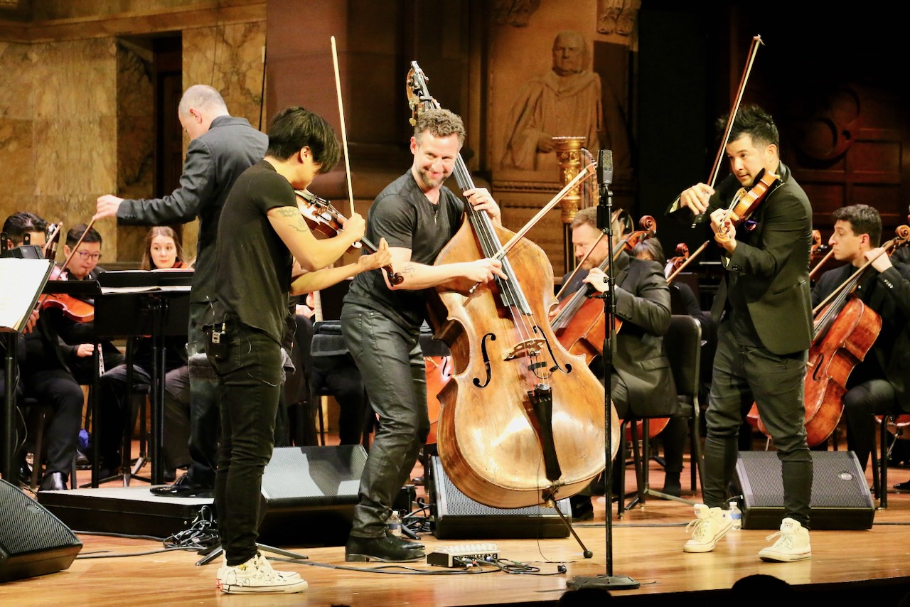 Two violinists flanking a double bassist during a performance onstage with a multiple cellists, a violist, oboist, conductor, and harpist visible behind.