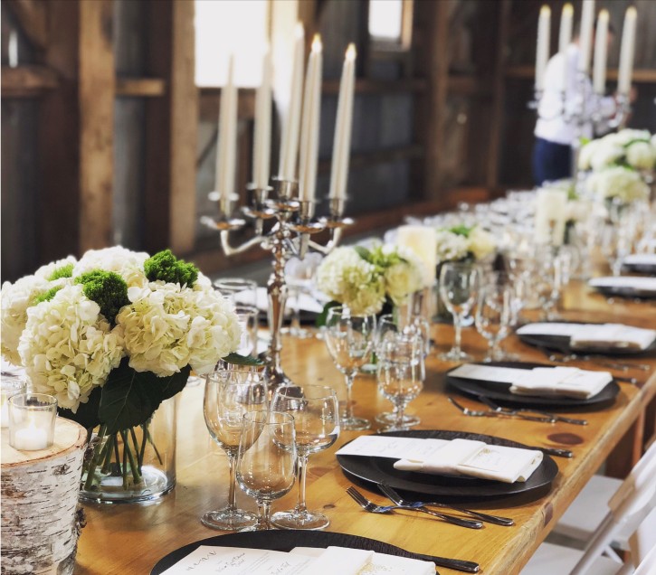 A table set for a dinner, with flowers and a candelabra. The supports of a barn are visible in the background.