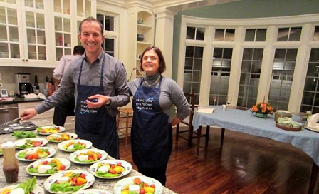 Maestro Rossen Milanov and former PSO Executive Director Melanie Clarke standing in a kitchen. They are both wearing aprons that read Princeton Symphony Orchestra. In front of them are a number of plates filled with food.