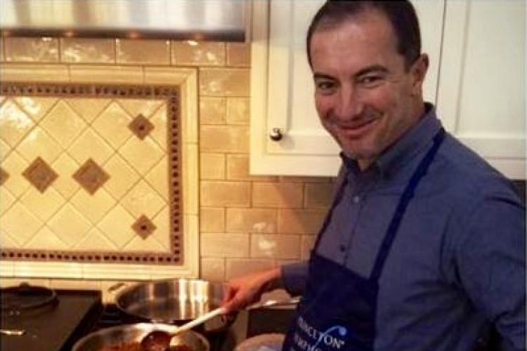 Rossen Milanov wearing a PSO apron, smiling at the camera, and cooking food in a pan on the stove