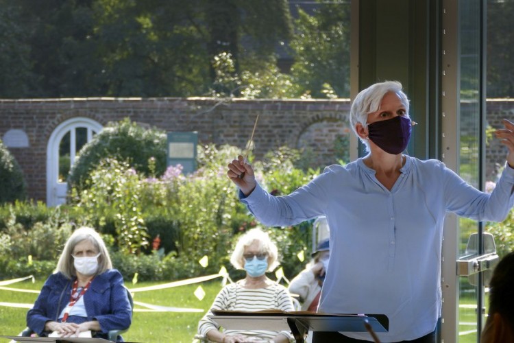 Nell Flanders, wearing a mask, conducts the orchestra. Visible behind her are two audience members, also wearing masks, and seated on the lawn.