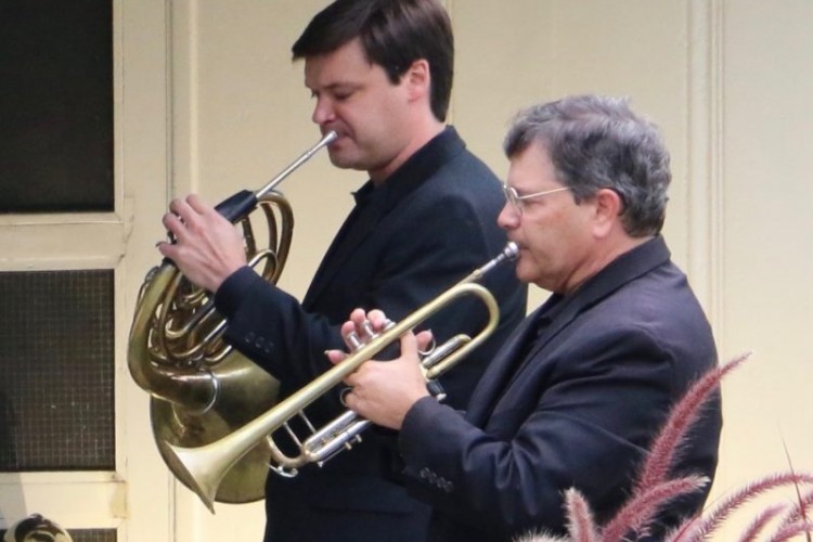 Two men standing next to each other. One plays the horn and the other plays the trumpet.