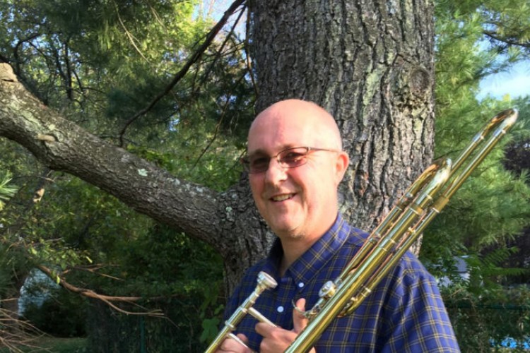 Lars Wendt stands in front of a tree holding a trombone and smiling