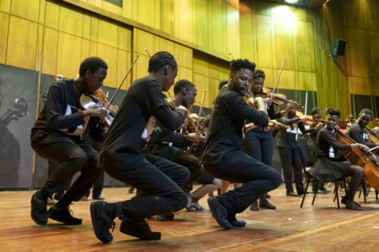 Violinists in the Buskaid Soweto String Ensemble performing, viewed in profile. Other performers with string instruments are visible in the background.