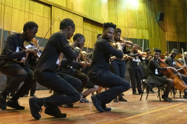 Violinists in the Buskaid Soweto String Ensemble performing, viewed in profile. Other performers with string instruments are visible in the background.