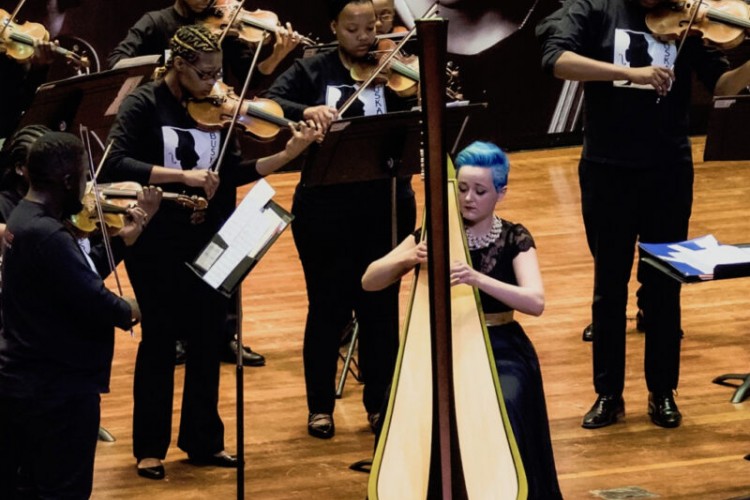 Jude Harpstar is seated and performing on the harp. Buskaid musicians playing violins stand behind and beside her.