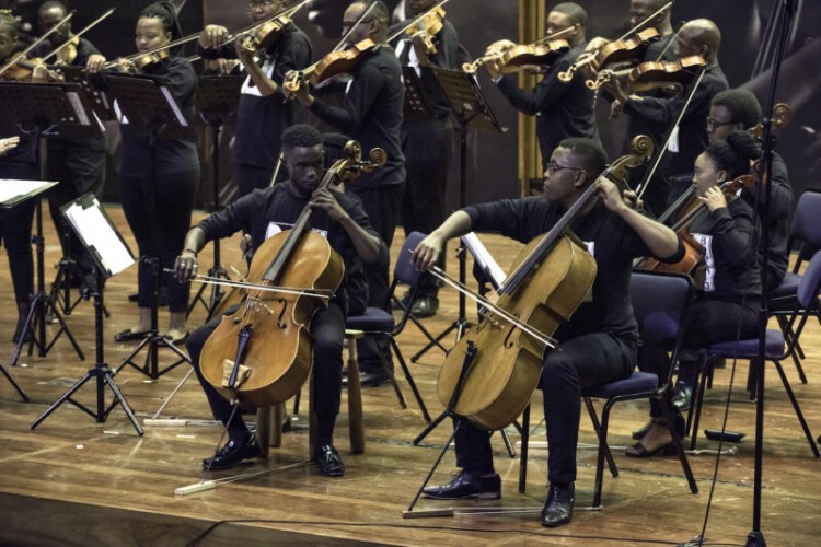 Two cellists seated and playing at the front of the stage, facing the audience. Behind them, more cellists and a group of standing violinists are also playing