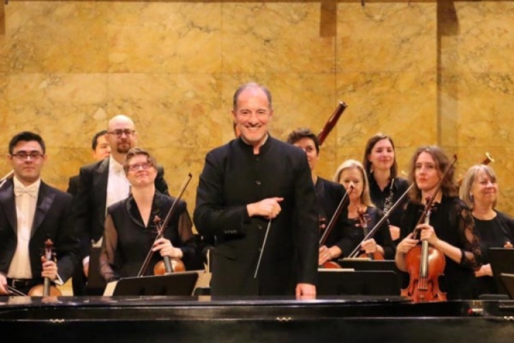 Edward T. Cone Music Director Rossen Milanov on stage, in front of the orchestra after a performance. He is facing the camera and smiling, holding his baton to his chest. Behind him, the musicians are also smiling and holding their instruments. 