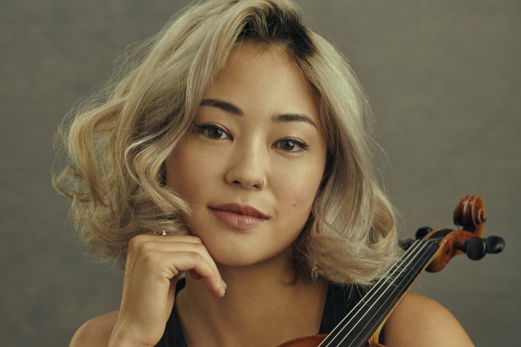 Simone Porter leans her chin on one hand and cradles her violin with the other while smiling at the camera