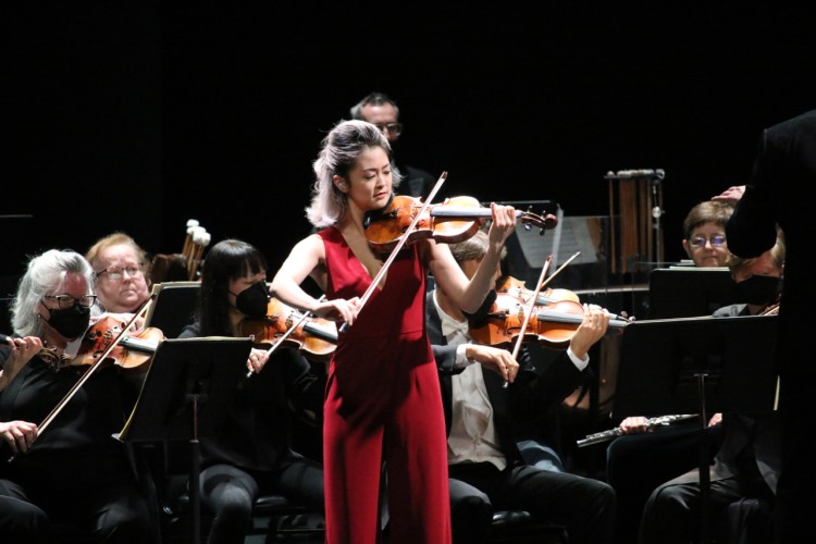 Simone Porter plays the violin. Behind her are the seated members of the orchestra, also currently playing.