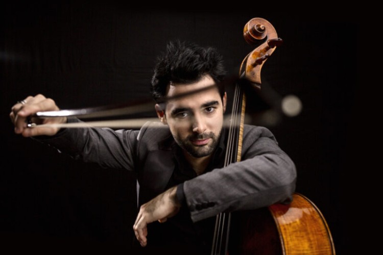 Pablo Ferrandez facing the camera with his left arm hooked around the fingerboard of his cello and holding the bow towards the camera with his right hand