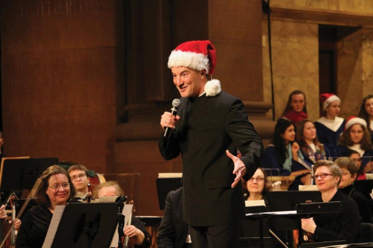 Rossen Milanov stands onstage holding a microphone and wearing a Santa hat. Behind him are members of the orchestra and members of a choir, also wearing Santa hats