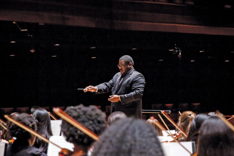 Conductor Kenneth Bean stands in front of an orchestra, holding a conductor's baton. 