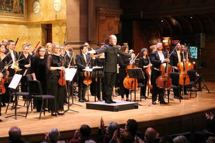 Music Director Rossen Milanov stands facing the audience with his arms open. Behind him the orchestra is standing, holding their instruments and facing the audience. The audience is visible in the foreground applauding.