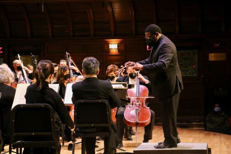Conductor Kenneth Bean stands in front of the orchestra, conducting. Bean and the orchestra are seen in profile, so the orchestra is on the left hand side of the picture, and Bean on the right.