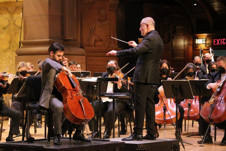 Cellist Pablo Ferrandez is seated to the left of the image, playing the cello. To his right is Rossen Milanov, standing and conducting the orchestra, which is visible behind both Rossen and Pablo. 