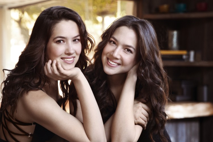 Christina and Michelle Naughton smile at the camera. They both are resting their heads in the palm of their hands.