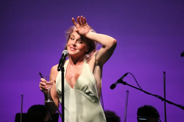 Singer Storm Large stands on stage in front of a microphone. She has one hand dramatically splayed on her forehead, and the other is holding a wineglass. Musicians and microphones are visible behind her against a purple backdrop.