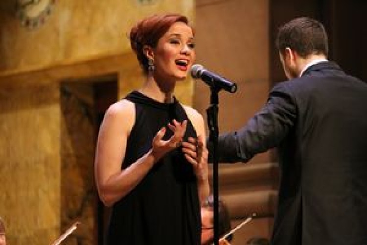 Singer Sierra Boggess stands in front of a microphone on stage. The conductor is visible behind her to the right.