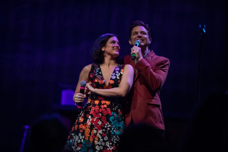 Vocal duo Alyssa Giannetti and Jason Forbach on stage. They are both holding microphones. Giannetti is smiling and looking up at Forbach while he sings