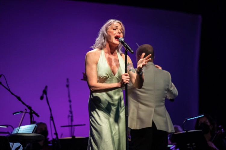 A close up shot of Storm Large on stage. She is holding onto a microphone stand and gesturing as she sings. Conductor Rossen Milanov is just visible behind her as he conducts the orchestra.
