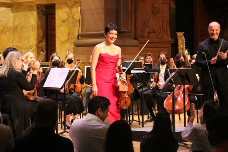 Violinist Anne Akiko Meyers stands to receive applause after her performance. Behind her, Rossen Milanov and the members of the orchestra are visible.