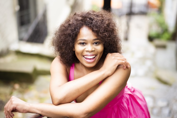 Soprano Pretty Yende is smiling directly at the camera. She is standing on a staircase and leaning on the railing.