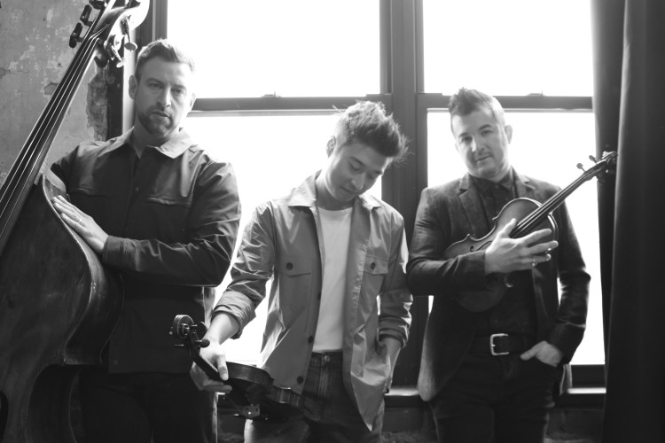 The members of trio Time For Three standing in front of a window. They are holding their instruments. The men on the left and right are looking at the camera, but the man in the middle is looking down. The image is black and white.