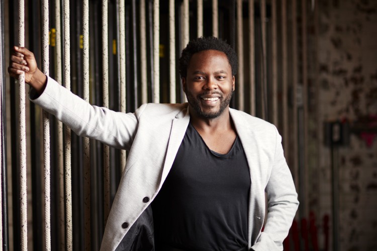 Baritone Will Liverman smiling at the camera. He is standing in front of a wall of ropes, holding onto one with his right hand. His other hand is in his front pocket.