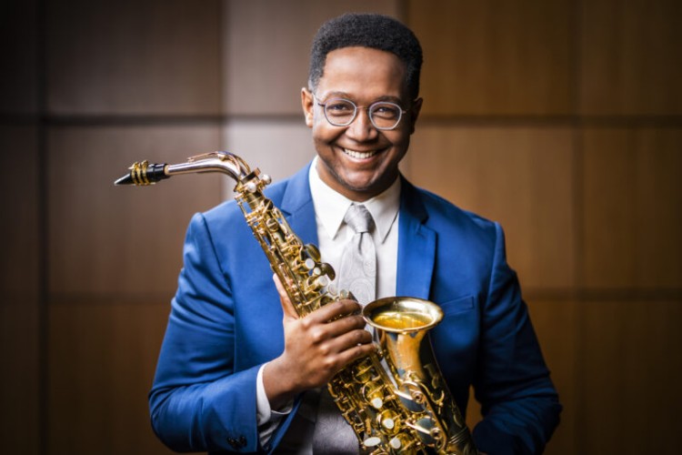 Saxophonist Steven Banks looks at the camera and smiles. He is holding his saxophone. The image is from the waist up.