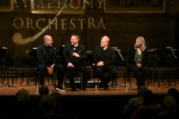 Three men and one woman sit on a stage with microphones as they engage in a conversation in front of an audience. 