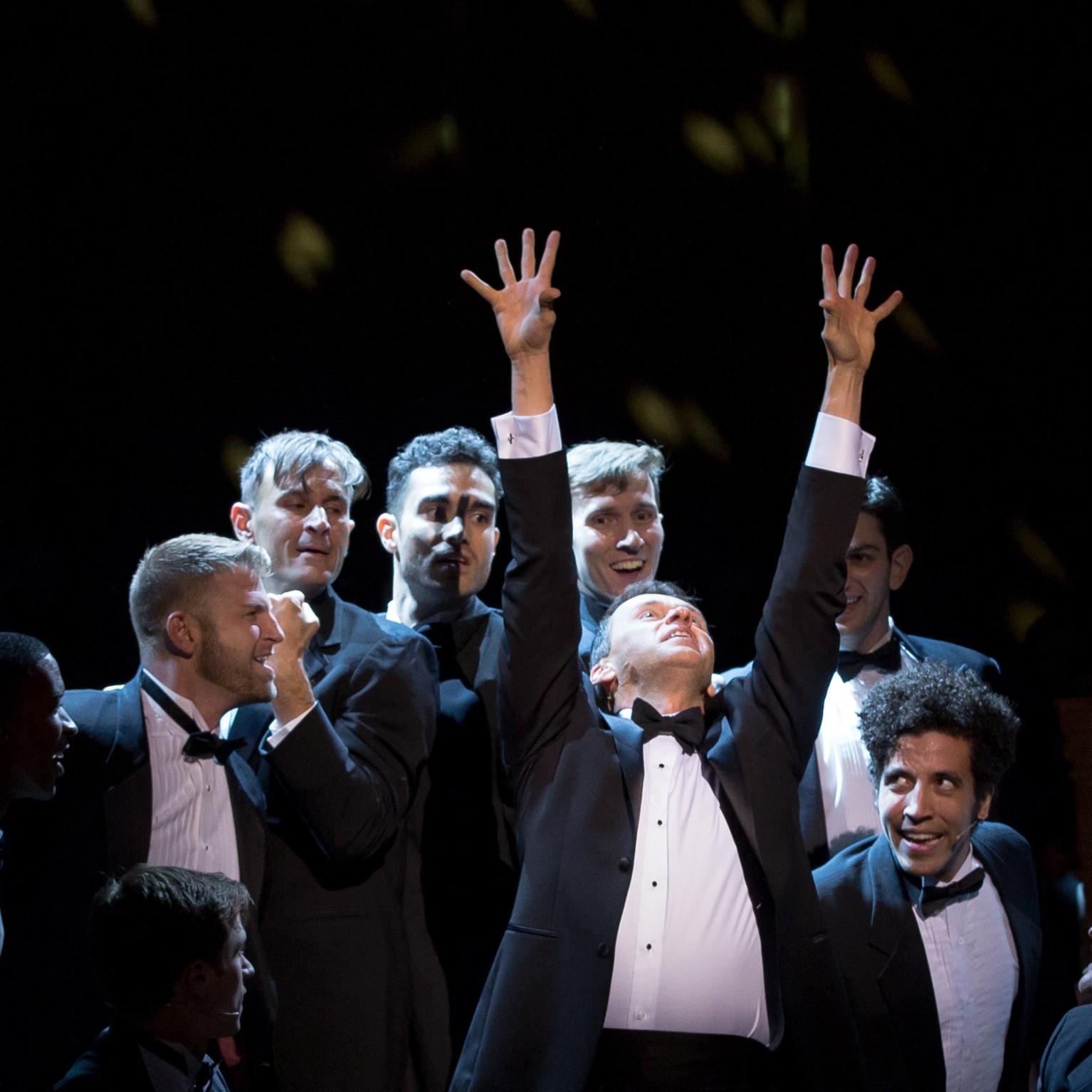 Men in black tuxedos rejoicing on stage around a man with hands raised up high