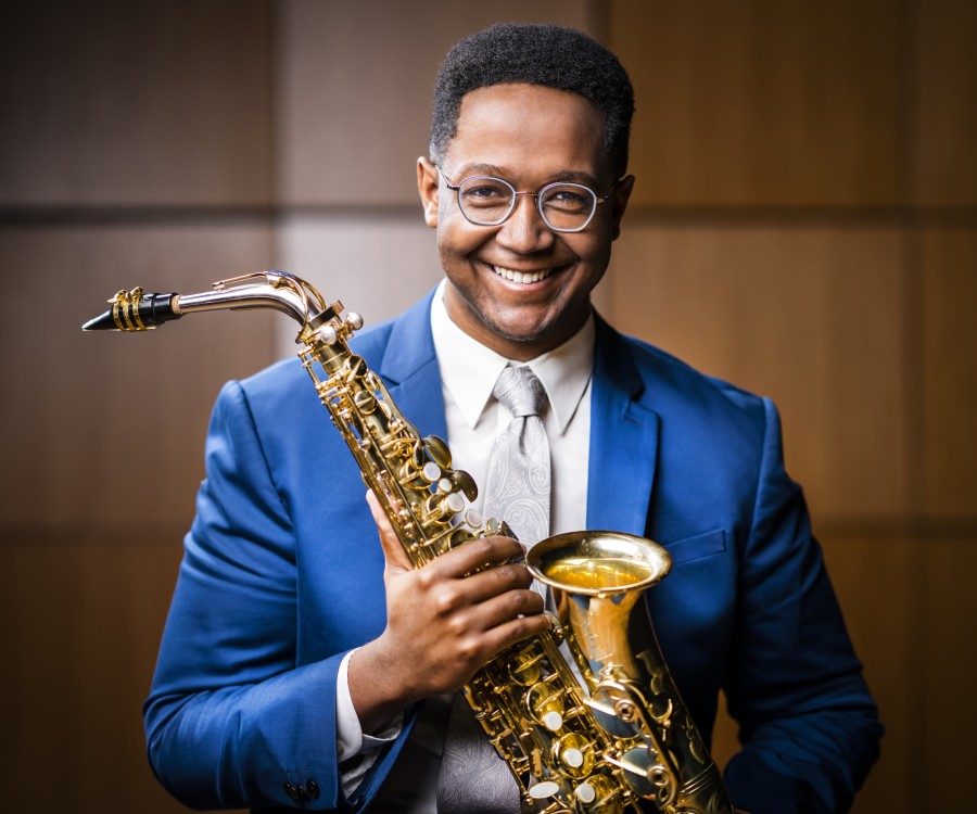 A smiling Steven Banks wearing a bright blue suit and holding up his saxophone in front of a wood wall