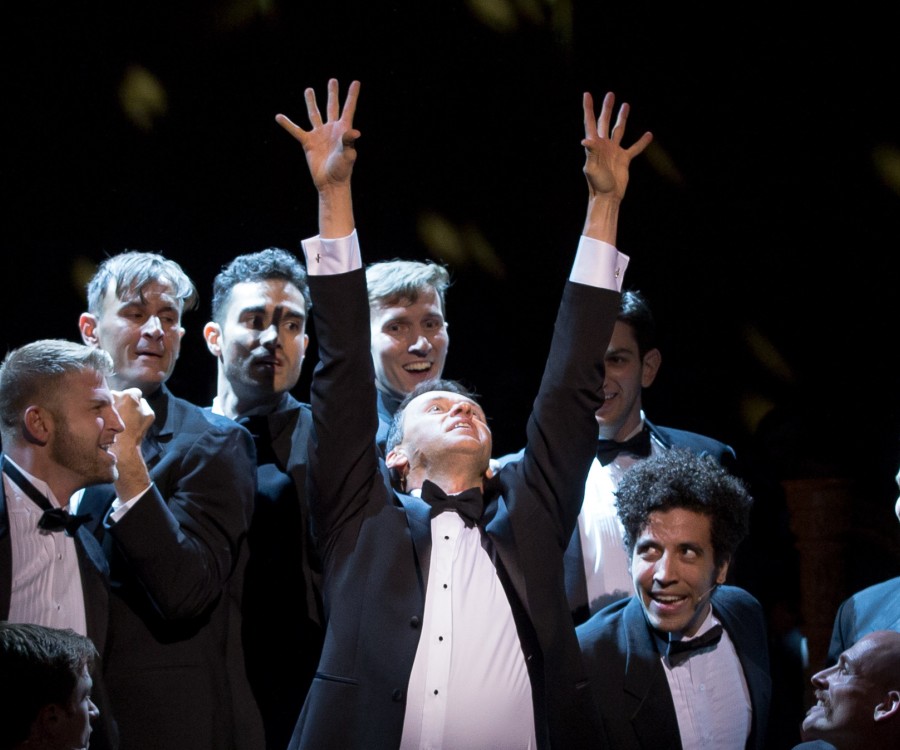 Male singers in tuxedos surround a central, spotlighted figure also wearing a tuxedo, with hands raised up towards the heavens