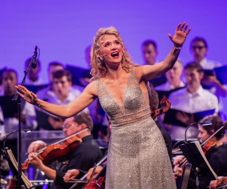 Vocalist in sparkling gown singing joyfully with outstreched arms in front of an all-male choir with orchestra members in between.