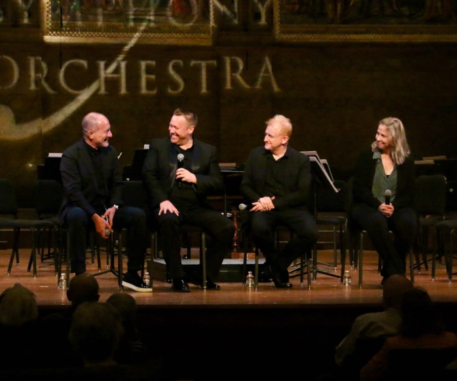 Three men and one woman sit on a stage with microphones as they converse in front of an audience. 