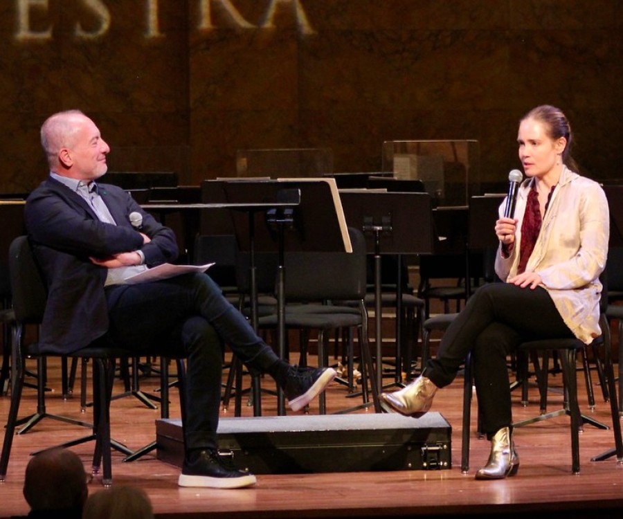 Two people sit on a stage with microphones, both holding microphones and in mid-conversation. 